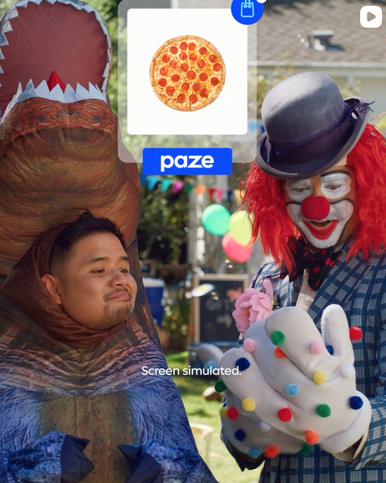 A screenshot of a video where a man dressed as a clown is using his phone to order a pizza with Paze while a man in a t-rex costume watches.